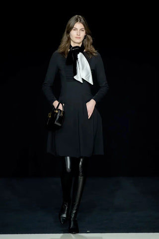 Model in runway in a long-sleeves black midi dress with thigh-high boots and a black and white scarf tied around her neck