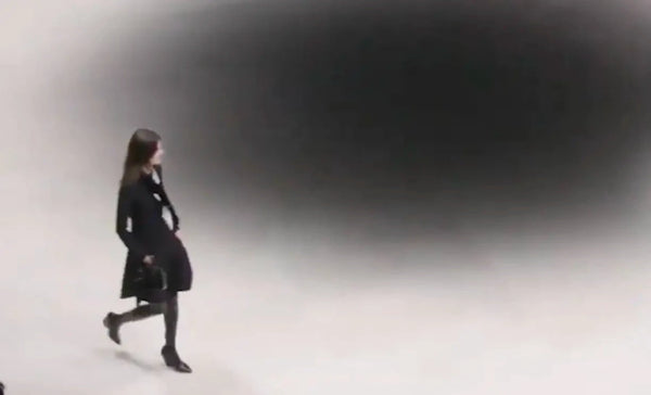 Model in runway wearing long-sleeve black midi dress with thigh-high boots and a black and white scarf tied around her neck