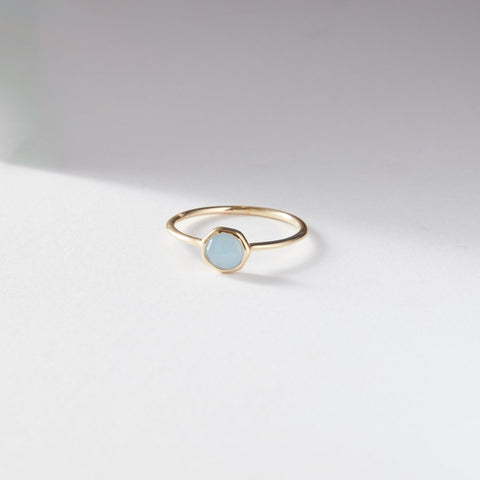 The aquamarine mini mood ring from bluboho, crafted in 10k yellow gold and featuring a round pale blue aquamarine gemstone, is displayed on a white surface. The ring casts a soft shadow diagonally across the background, highlighting its minimalistic and elegant design.