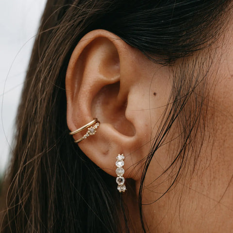 Close-up of a woman's ear adorned with multiple bluboho earrings. There is a Large Moonglade Hoop with a small star on the upper part of the ear, and a line of three diamonds in a drop earring dangling below. The woman's hair is dark and slightly damp against her skin.