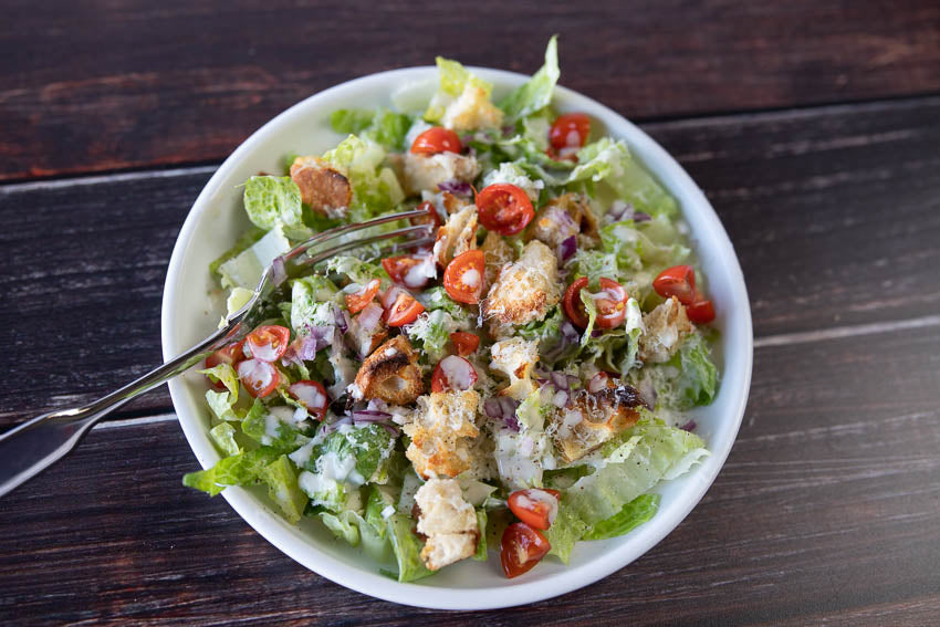 A large bowl with lettuces, diced tomatoes and finely chopped onions. With homemade croutons and a white creamy sauce drizzled on top  