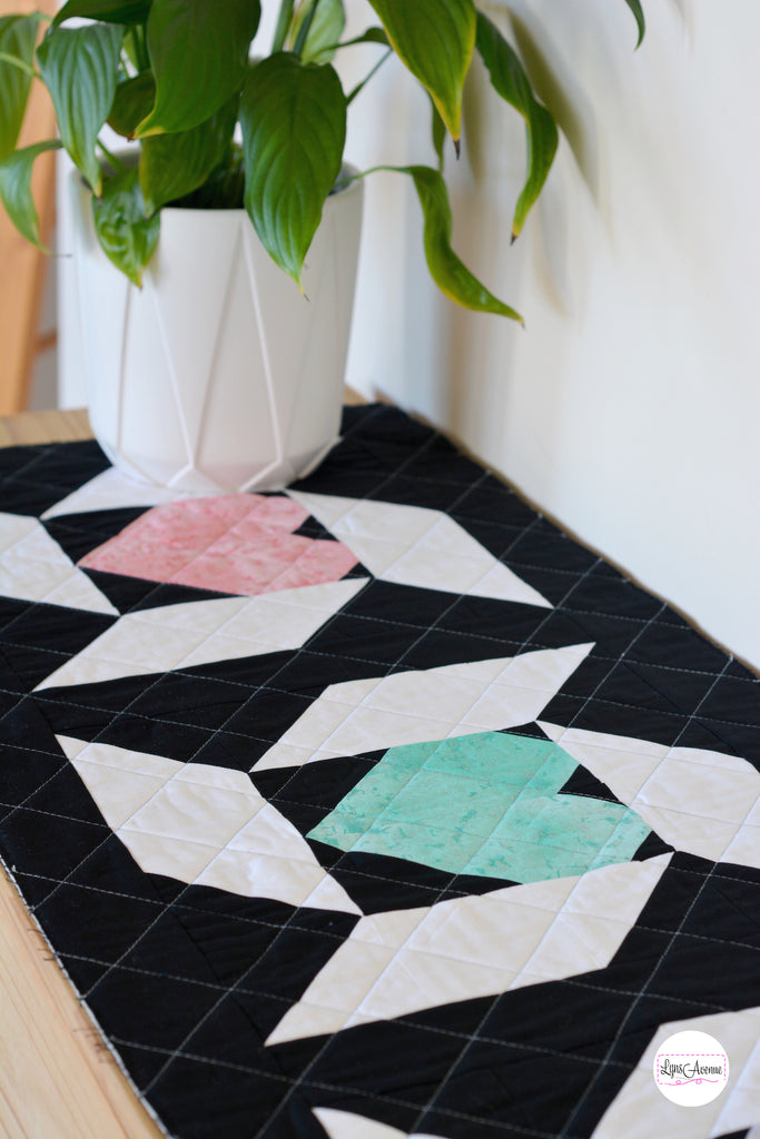 Picture of a table runner in black and white colours for the background and pink and green hearts. A white pot with a green plant is sitting on top.