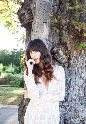 Bride leaning against tree, folded arms, wearing low front beaded lace wedding dress.