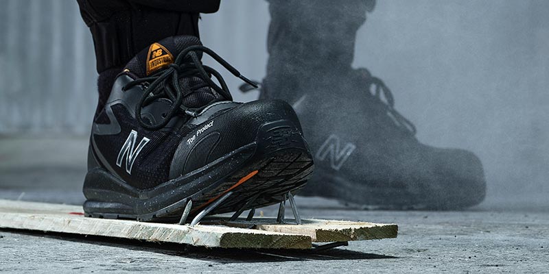 A person wearing a pair of black/orange New Balance Logic safety shoes, standing on wooden planks with sharp nails poking out of them showcasing the anti-penetration sole of the safety shoe