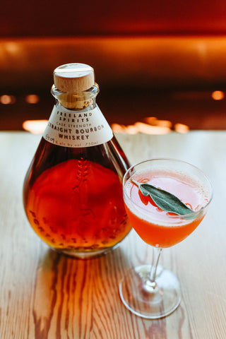 Freeland Cask Strength Bourbon bottle and Paper Airplane cocktail garnished with sage on a wooden table 