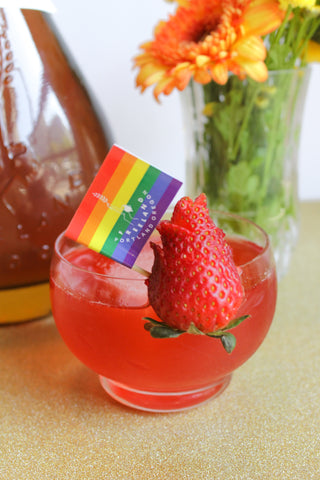 Freeland Spirits cocktail Strawberry Blvd garnished with strawberry and Pride flag. Set next to Freeland Bourbon bottle and flowers. 