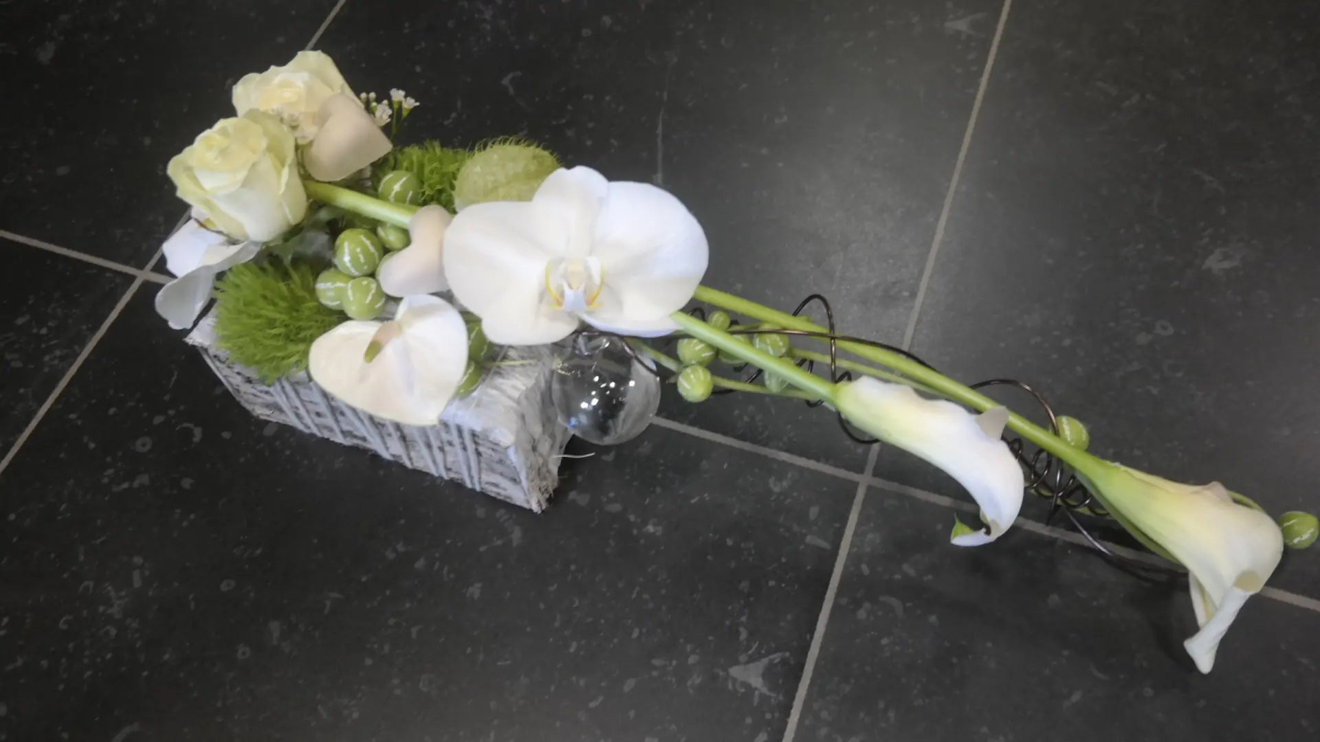 Witte bloemenschikking met calla, strakke groene bollen en mos, langs een kabelachtige vertakking op een donker betegelde vloer.