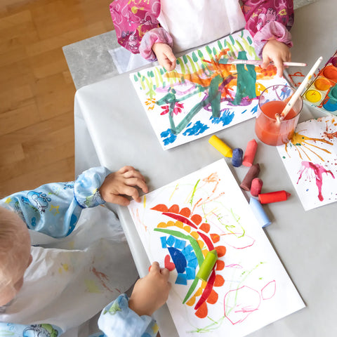 Zwei Kinder malen mit Wachsmalstiften und Wasserfarben auf Bastelunterlage.