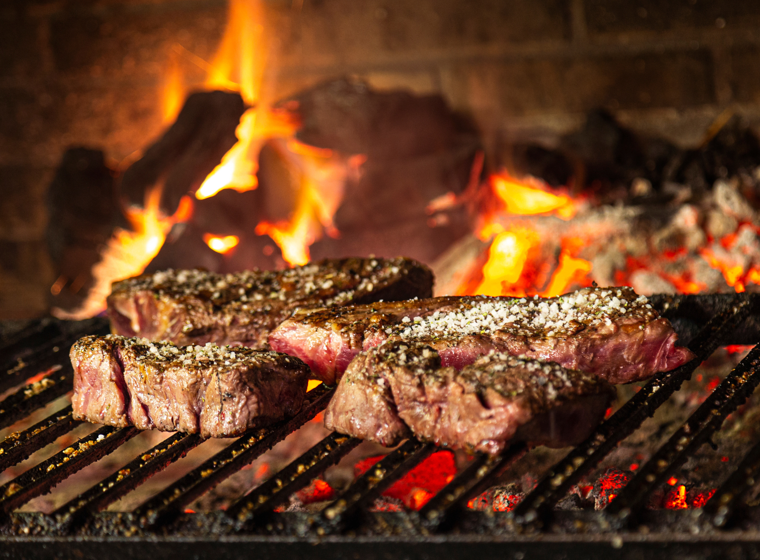 three steaks grilling over high flames and charcoal briquettes underneath