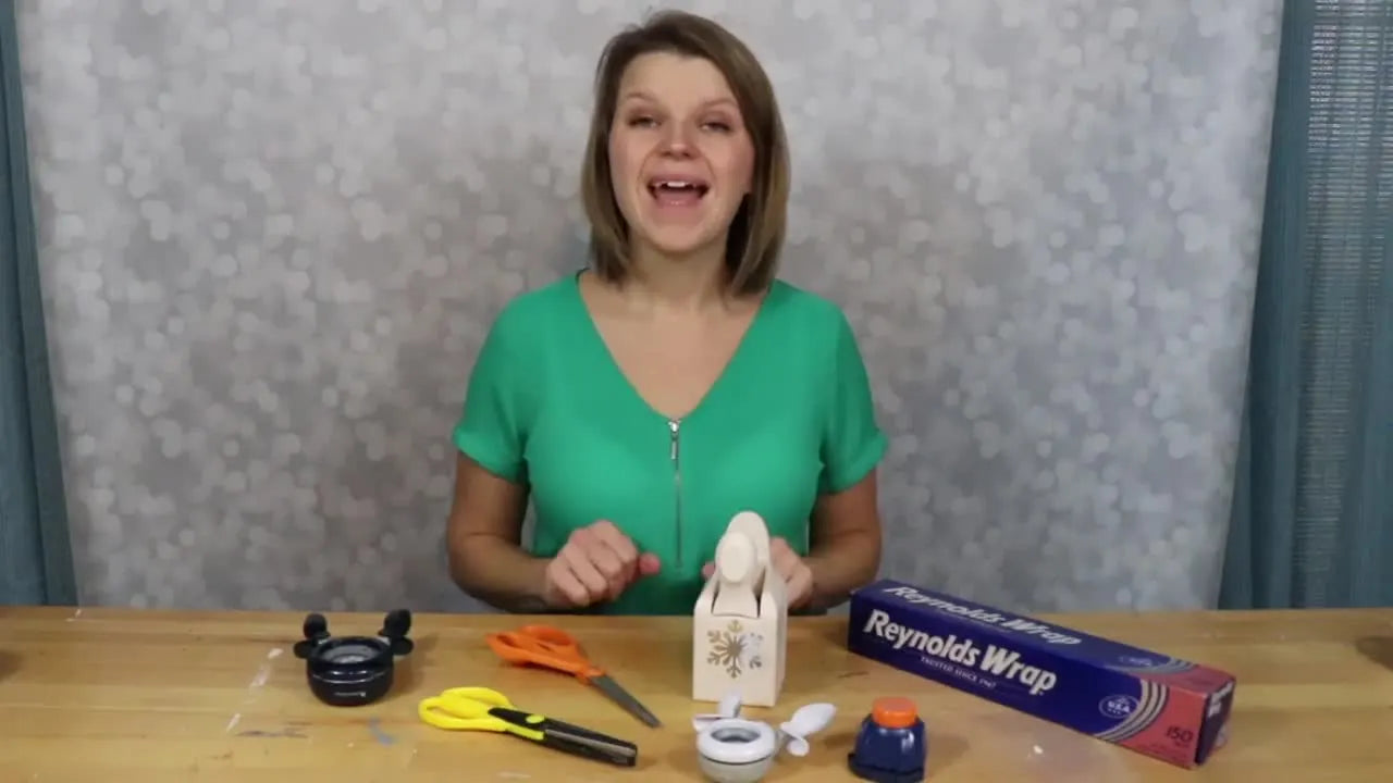 Presenter sitting at a table with various craft scissors, paper punches, and a box of aluminum foil.