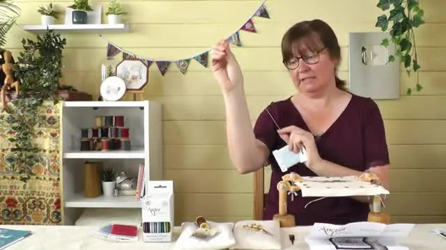 A woman measures a length of embroidery thread from her fingertip to her upper arm, just short of her elbow.