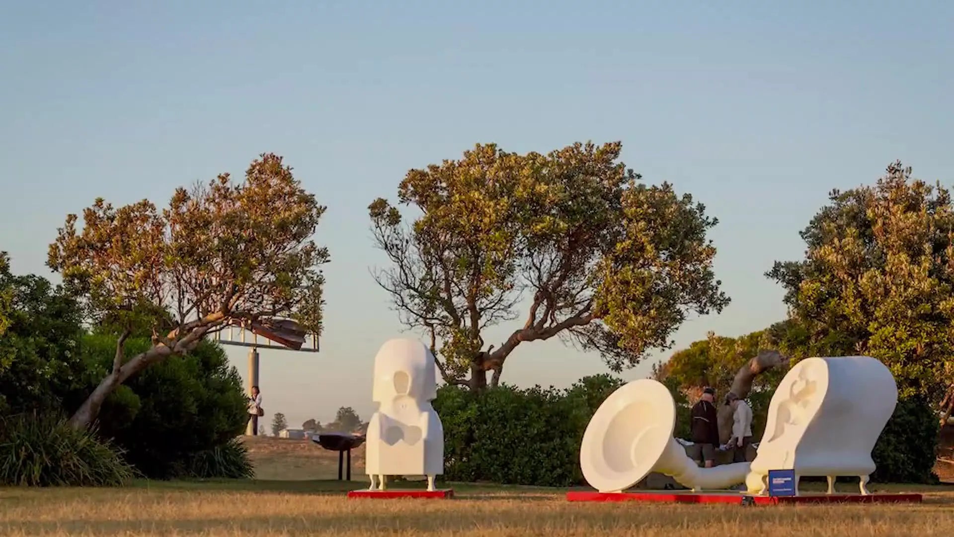 A large, white, abstract outdoor sculpture resembling a gramophone horn and a chair with a figure inside, set in a park-like environment.