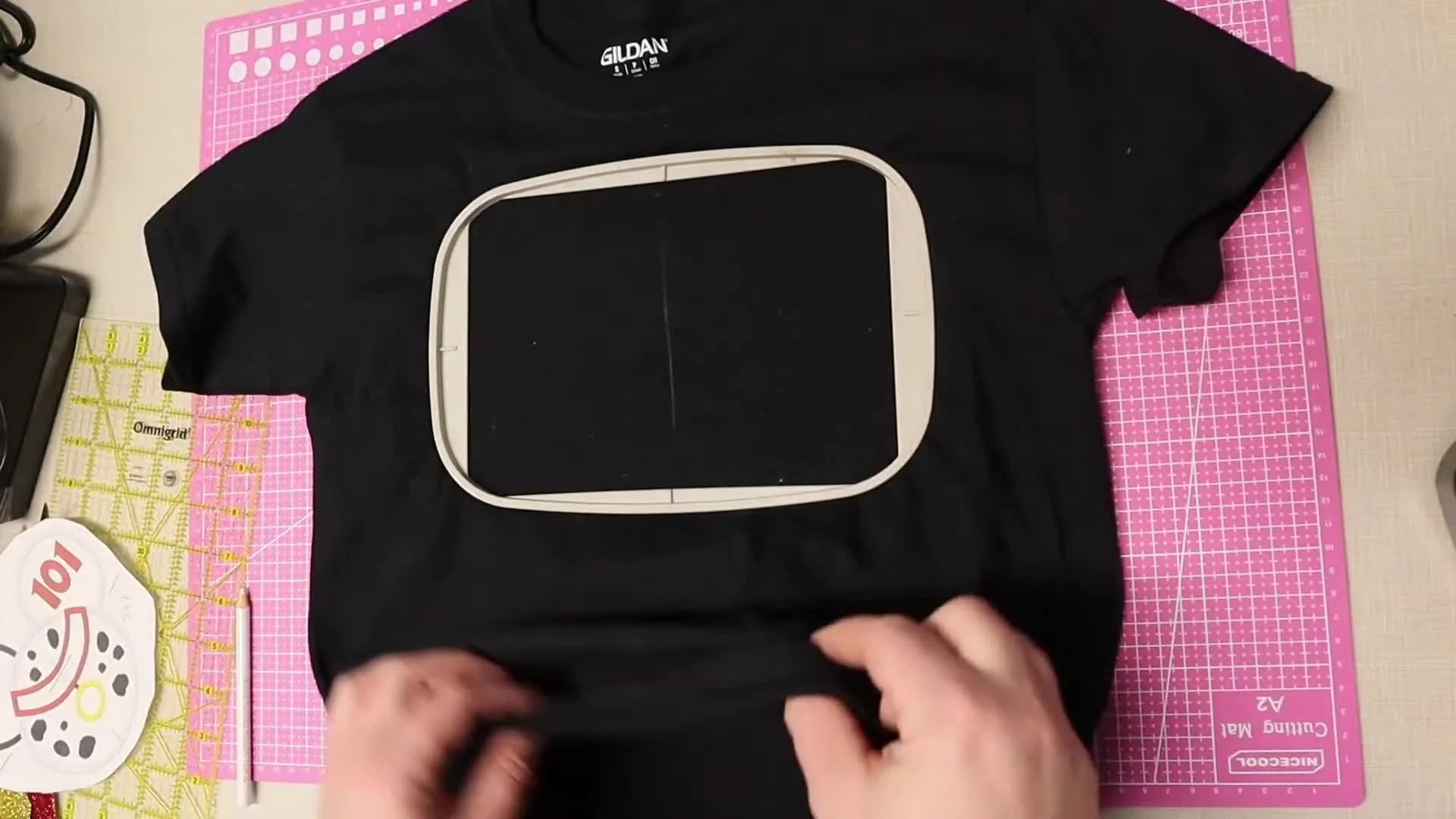 Hands tucking the back of the black t-shirt fabric away from the hooped embroidery area.
