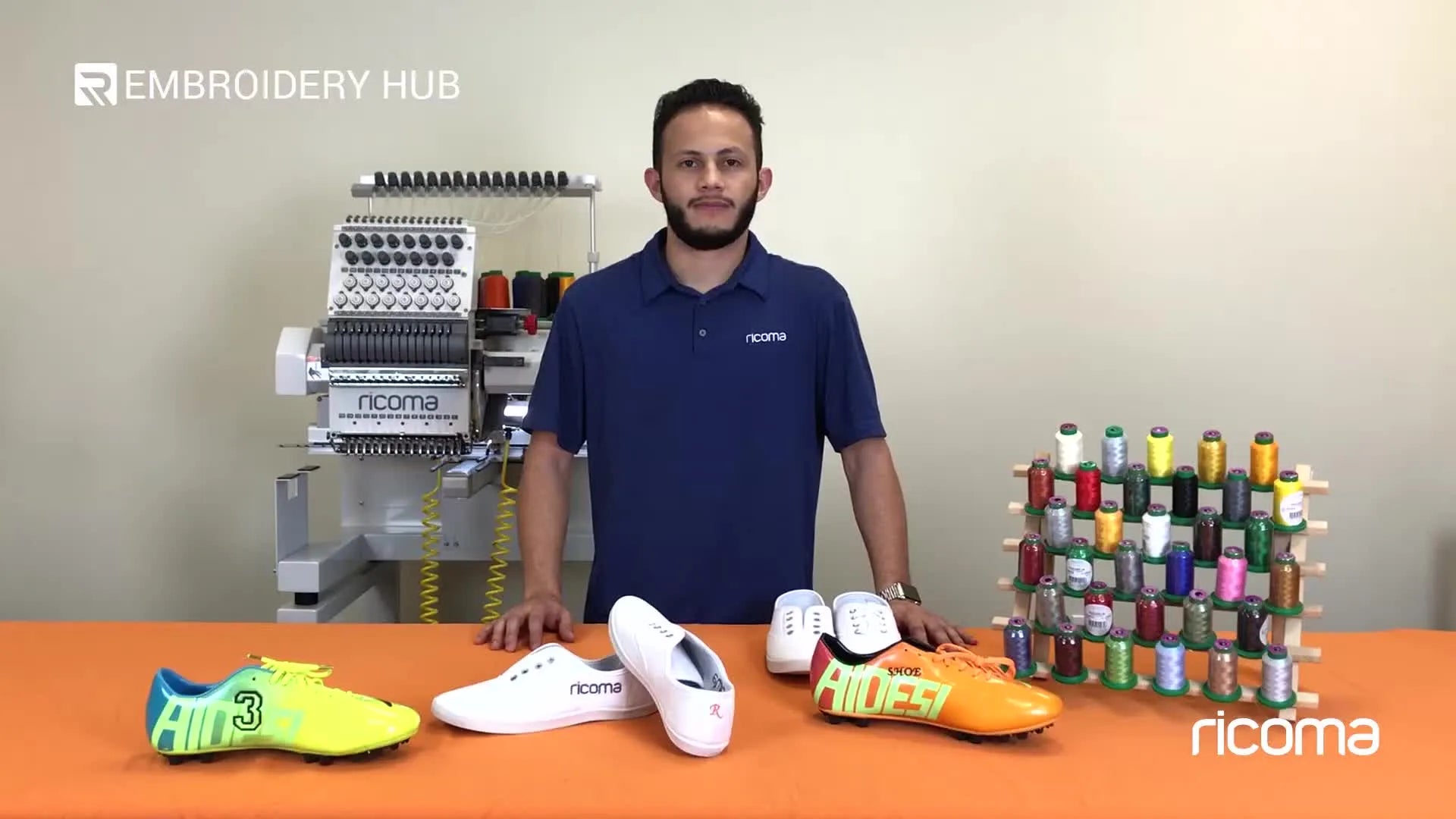 Man presenting embroidered shoes with embroidery machine in background.