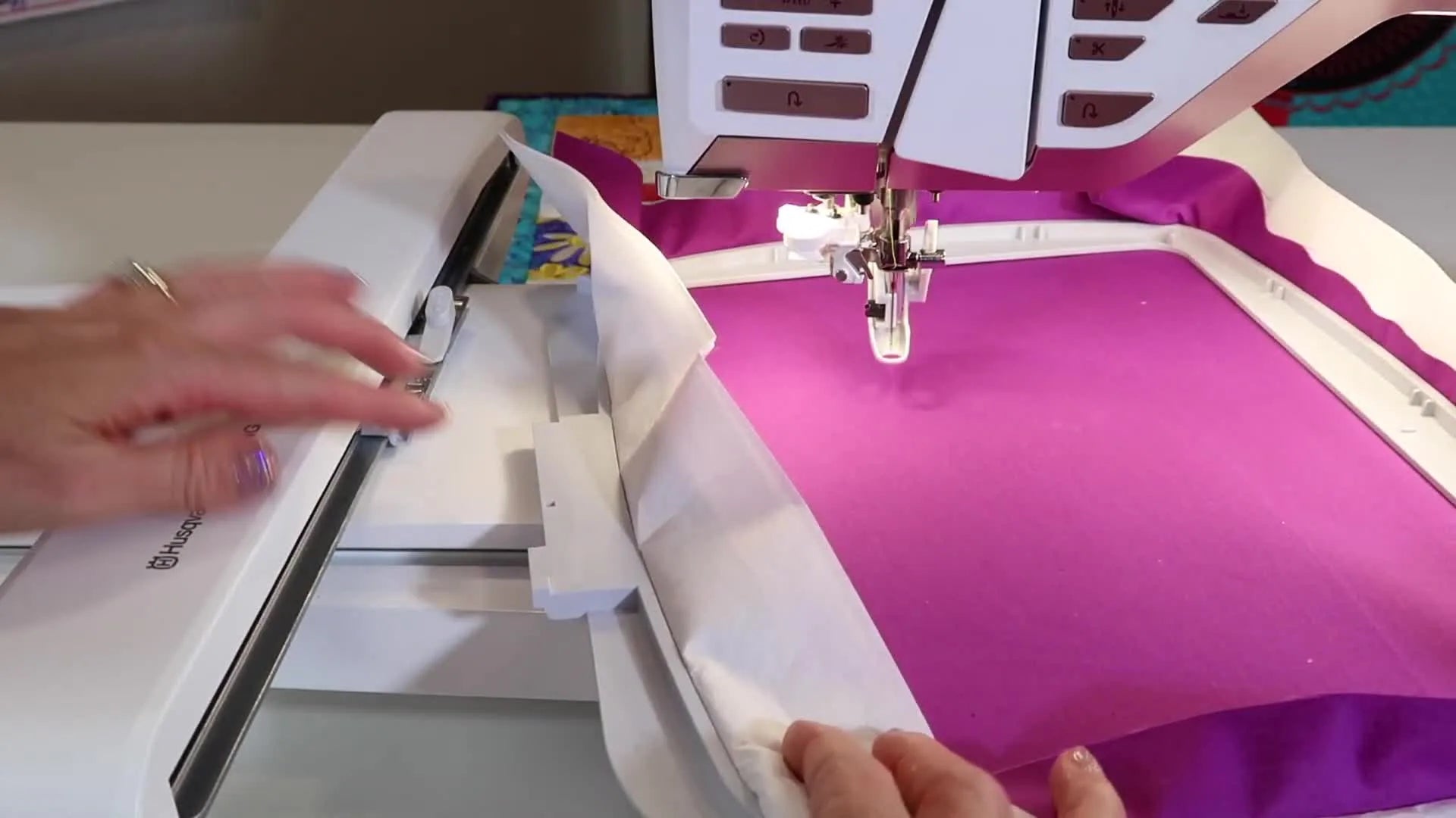 A hand sliding the hooped fabric's bracket into the embroidery arm attachment point, pointing to the silver top and gray lower parts.