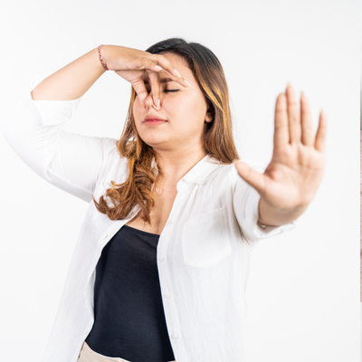 The image shows a person with their hands on their head. The person is a woman with long hair wearing a blouse. The background appears to be a wall.