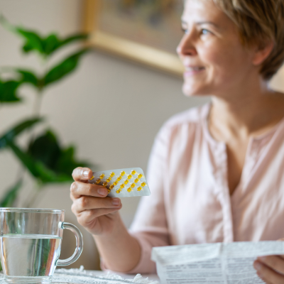 The image shows a woman holding up some pills while having a coffee indoors.
