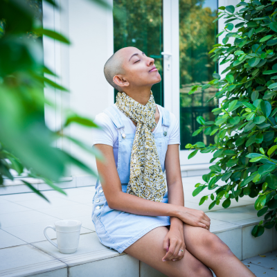 The image features a person sitting on a ledge outdoors. The individual is wearing fashionable clothing and accessories, with a tree and flowerpot visible in the background.