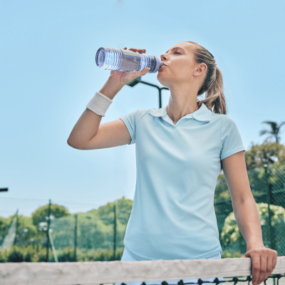 The image shows a woman outdoors, wearing clothing, drinking from a bottle against a sky background.