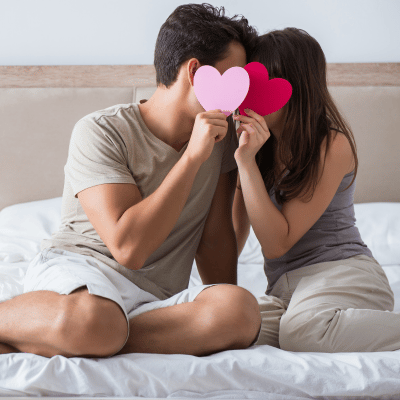 The image depicts a man and woman kissing on a bed both holding a heart shape paper covering their faces