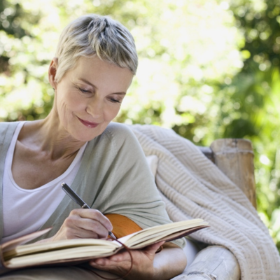 The image shows a woman sitting outdoors, writing in a book. She is focused on her task, surrounded by trees.