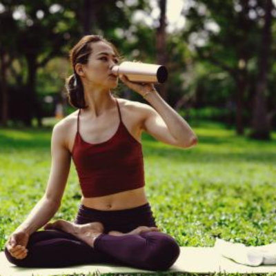 The image depicts a woman drinking from a bottle in an outdoor setting. She is surrounded by grass and trees, and appears to be engaged in physical fitness activities like yoga or playing frisbee in a park.