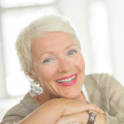 The image shows a woman with her arms crossed, smiling. She is indoors and is wearing a watch as a fashion accessory. The focus is on her facial features such as her smile, skin, eyebrows, and wrinkles.