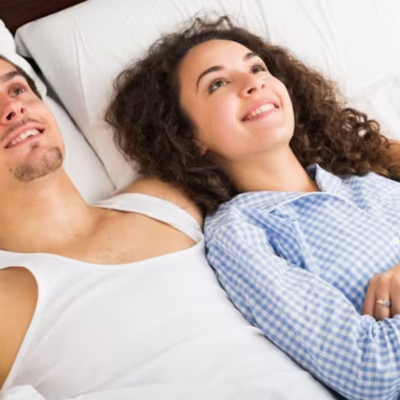 The image features a man and a woman lying in bed. Both individuals appear comfortable and are smiling. The setting is indoors against a wall.