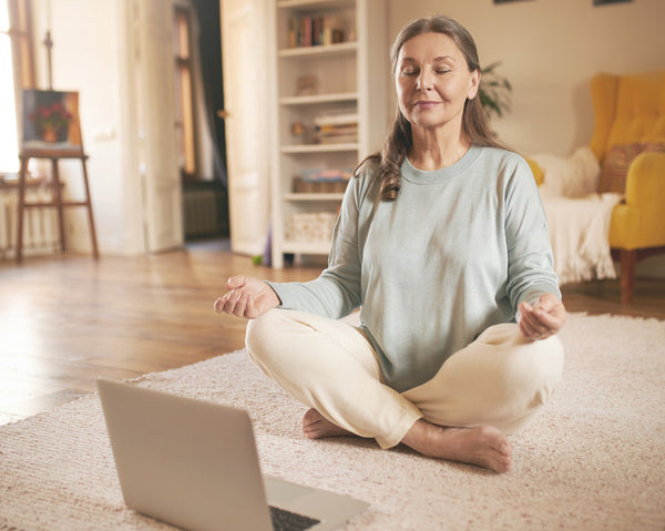 A woman sits cross legged in front of a laptop, meditating.
