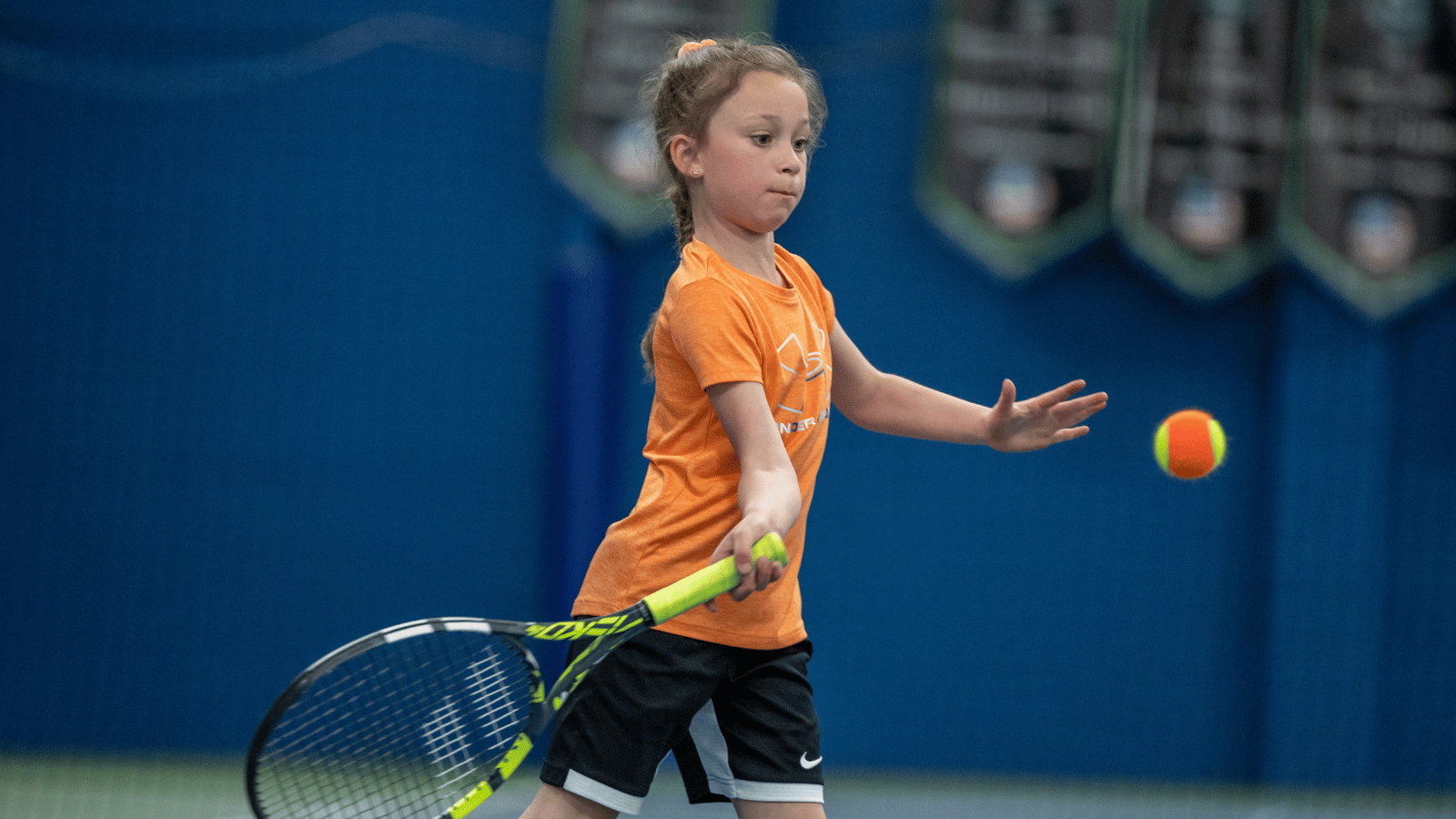 Young girl in orange shirt hits orange ball with racquet