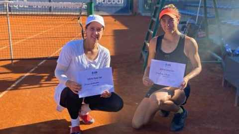 Two women pose on a red clay court with their UTR Pro Tennis Tour certificates.