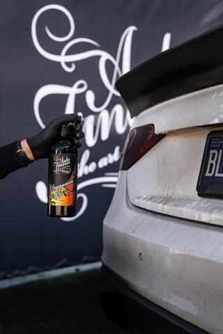 A bottle of Dynamite Traffic Film Remover being sprayed onto the trunk of a white vehicle covered in road grime.