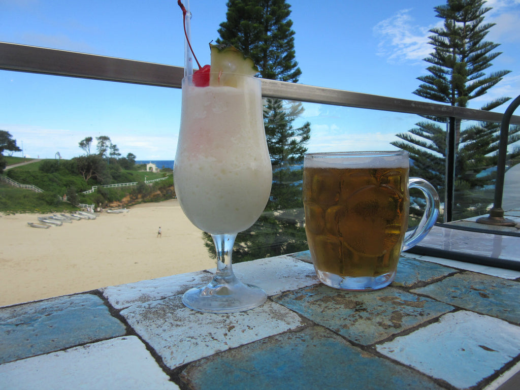 Two cocktails sit on a tiled wall of a rooftop bar, with a view of a sandy beach and grassy banks.