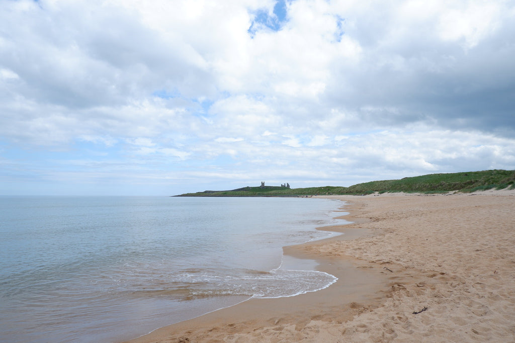Wide sandy beach with the outline of Dunstanburgh Castle perched on green clifftops.