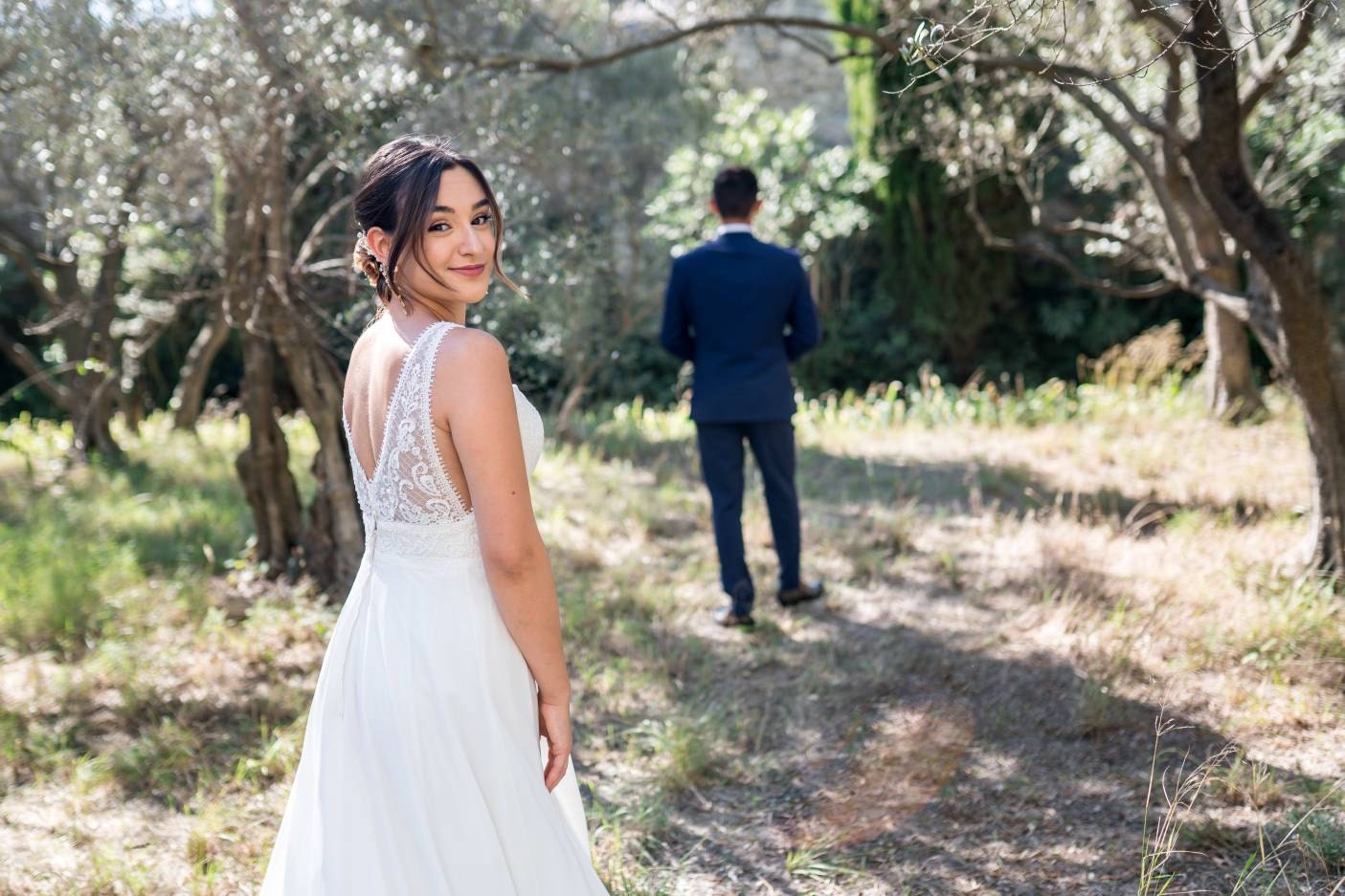 Mariée retournée vers nous dans un jardin provencal au milieu des oliviers avec le mari en arrière plan de dos