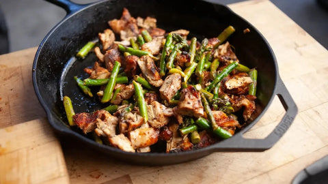 Cast iron deep pan with asparagus and chicken being mixed together, combining flavors for a hearty, flavorful dish.