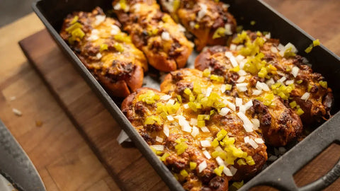 Downward view of a cast iron pan on a wooden table, filled with garlic bread chili dogs, showcasing a hearty, flavorful dish.