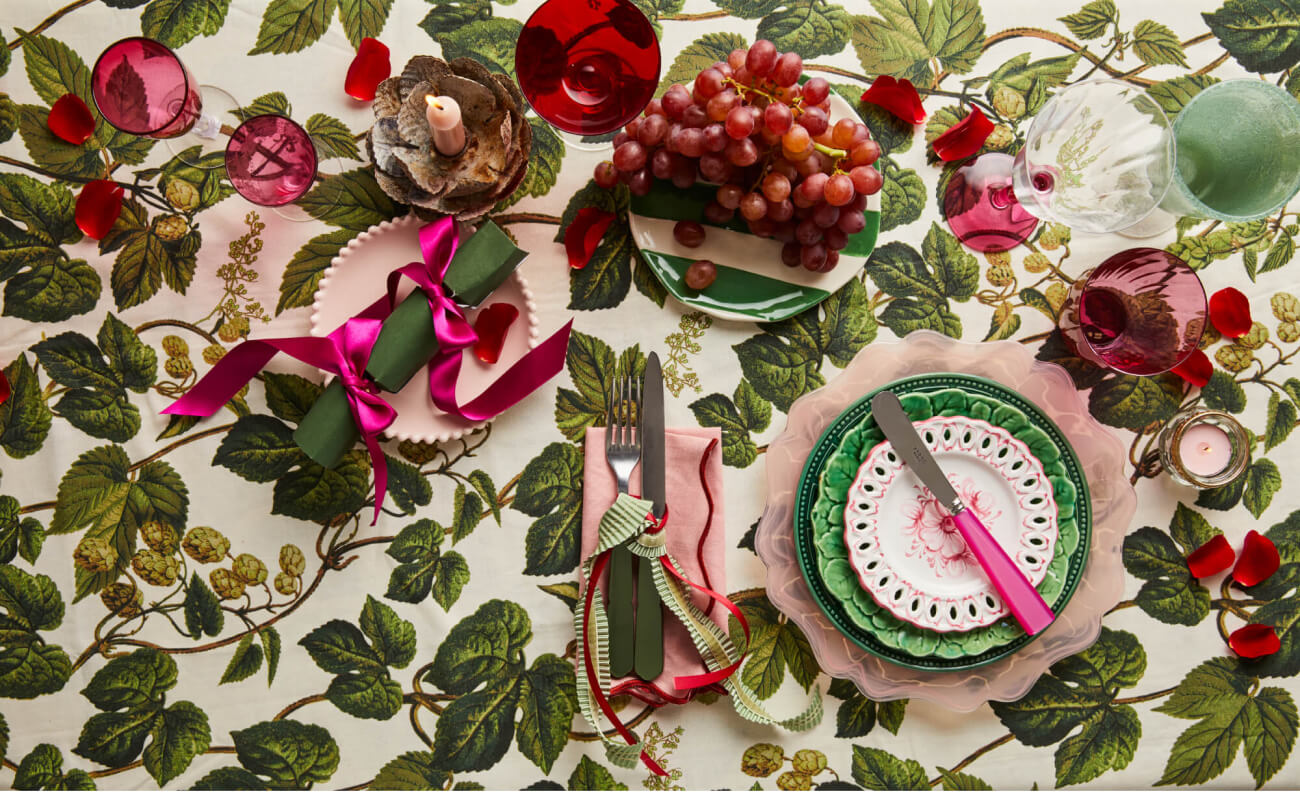 Christmas pink and green table setting from The Social Kitchen with green crackers with pink ribbon, mix and matched crockery, cutlery and glassware in pink and green, and fresh red grapes on vintage plates