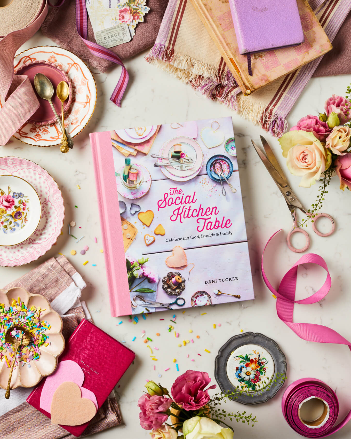 The Social Kitchen Table Cookbook on a table setting with pink, vintage plates, ribbons and craft scissors.