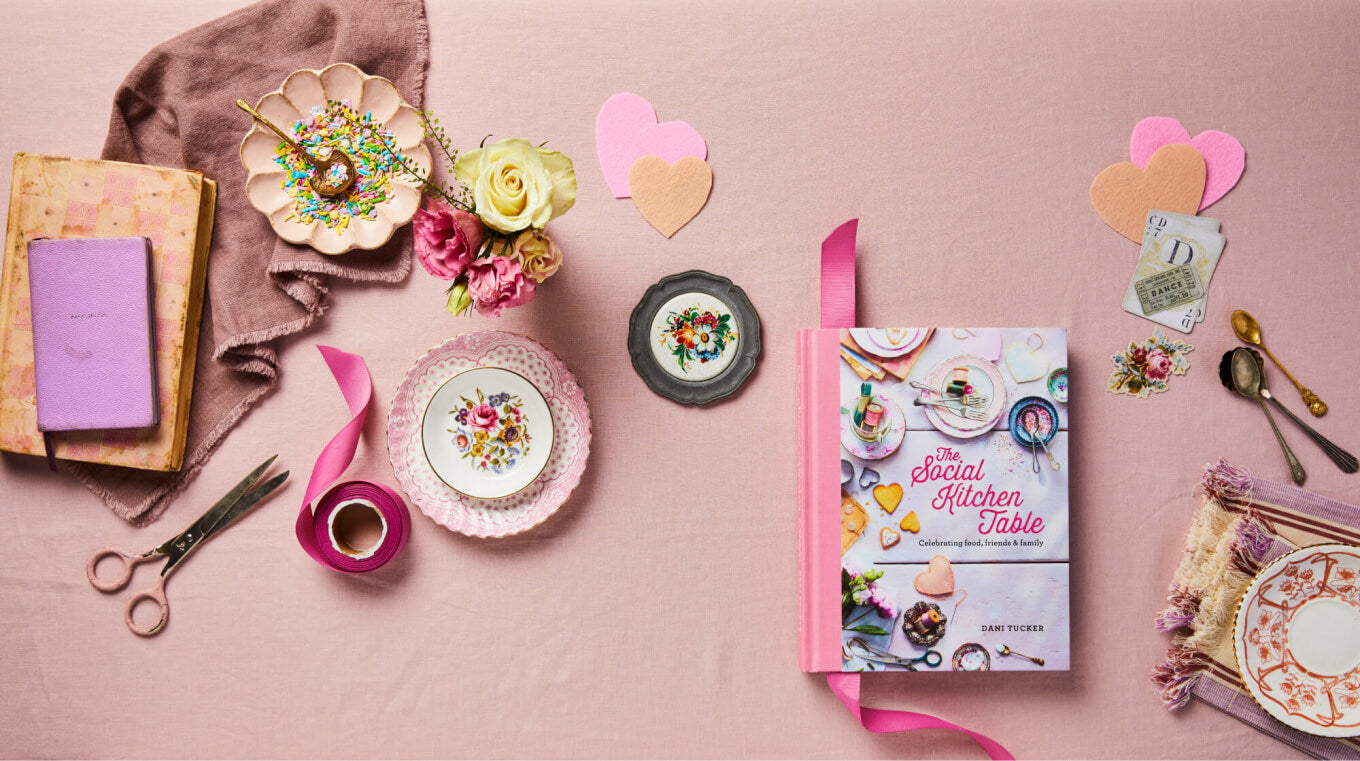 The Social Kitchen Table Cookbook on a pink background with vintage plates, flowers, craft scissors and journals.