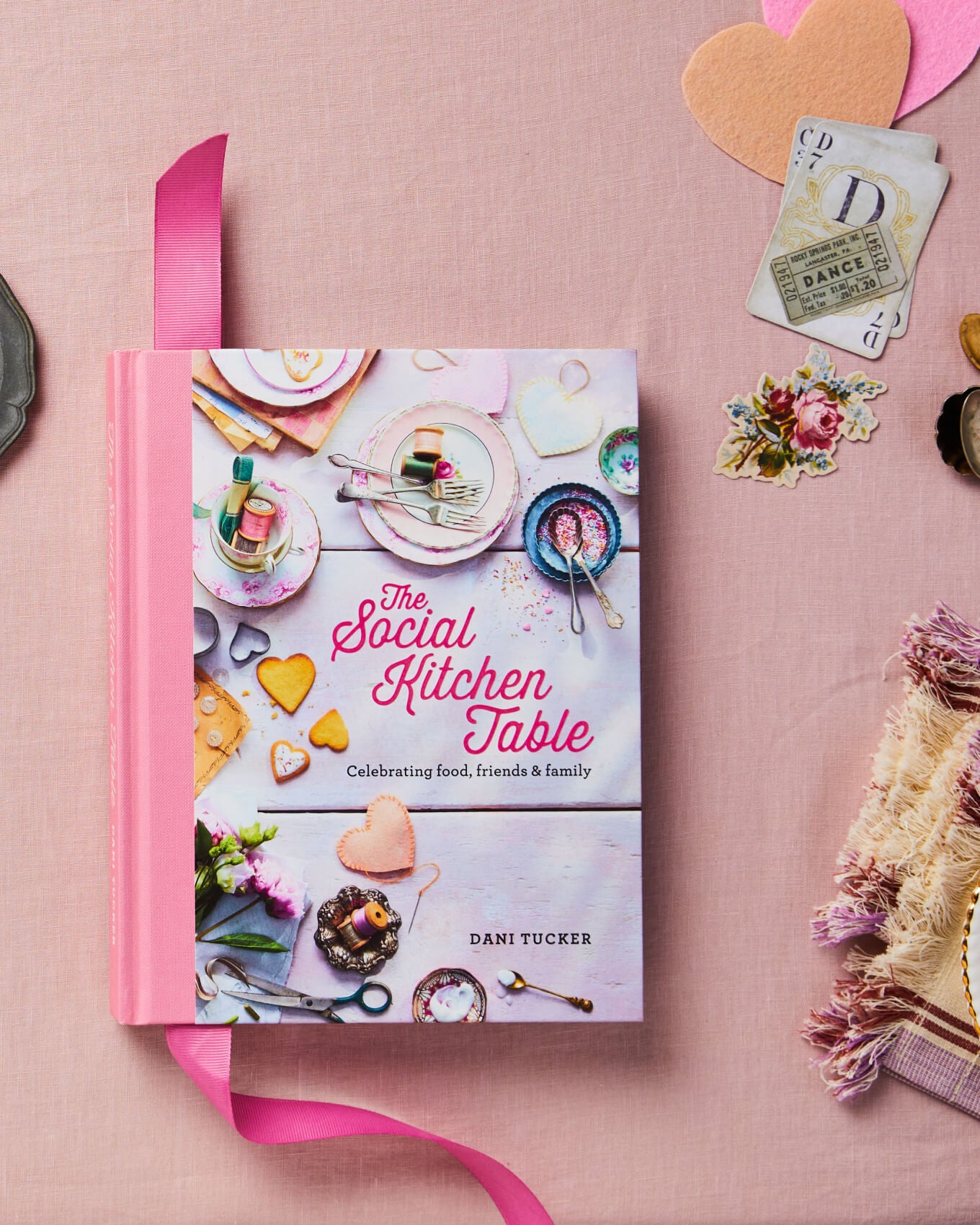 The Social Kitchen Table Cookbook on a pink background with craft items on the table