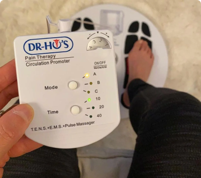 Person using a DR-HO'S Pain Therapy System with feet on white pads.