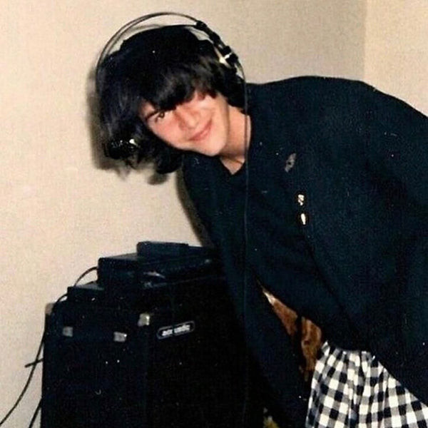 Keanu Reeves with large headphones, next to large speaker, smiling at camera, in black suit jacket and white/black plaid skirt.