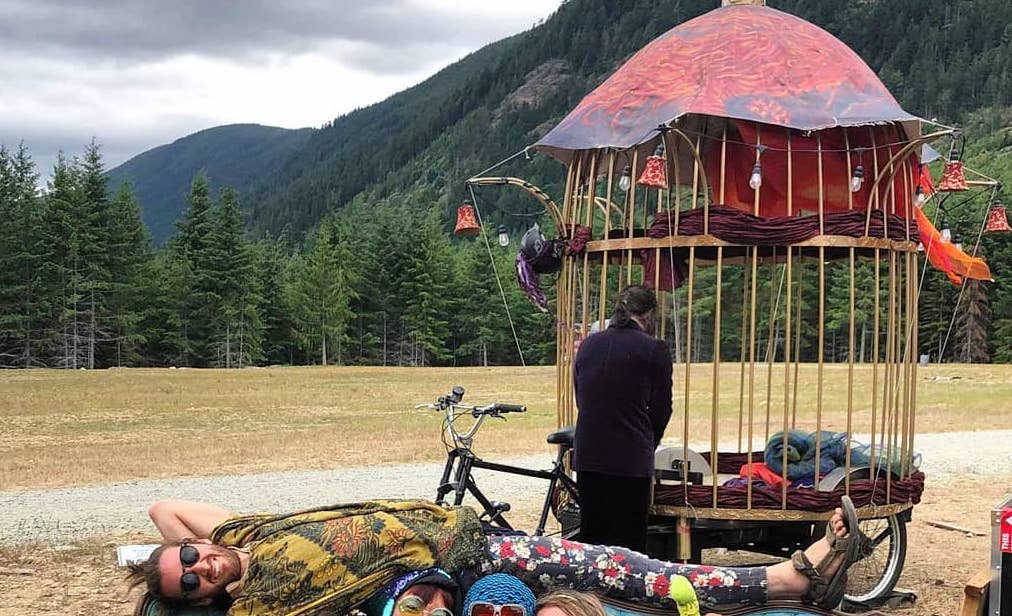 Me laying on back of a couch at Otherworld regional burn festival, with trees and mountain in background, giant bird cage bicycle behind, and others sitting on the couch.