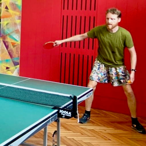 Male bodied person wearing a colourful skater skirt, and green t-shirt, playing ping pong, in front of a red background.