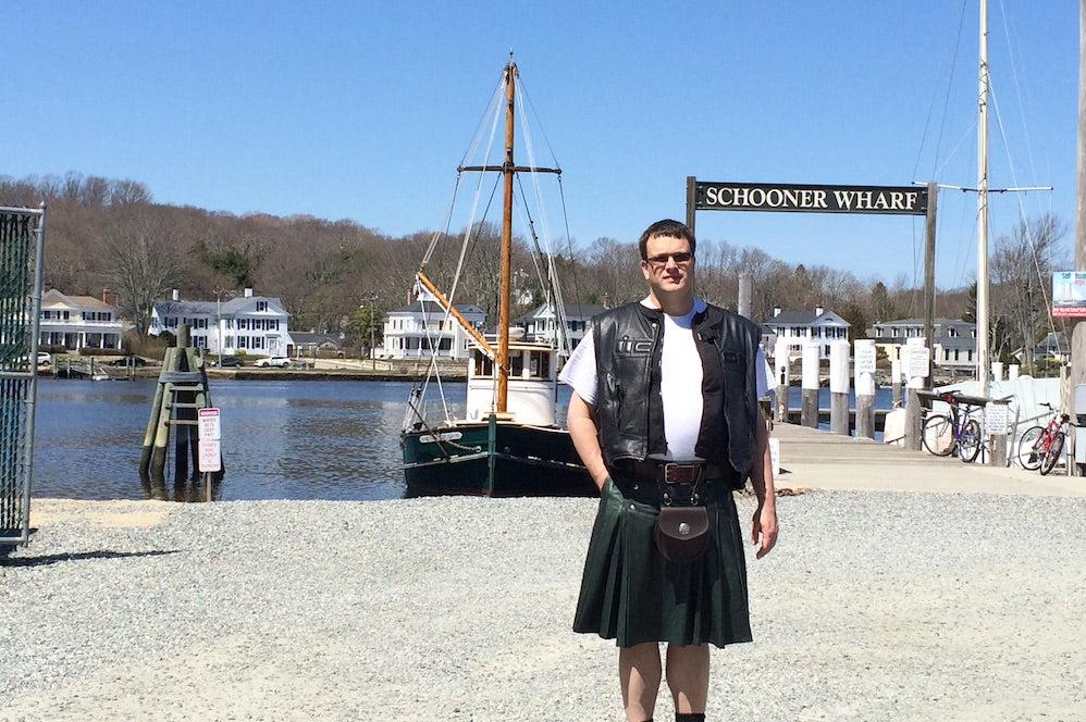 Murphy standing in front of 'Schooner Wharf' with sailboat in background. He has a leather kilt, and dark leather vest over a white t-shirt, and a sporran.