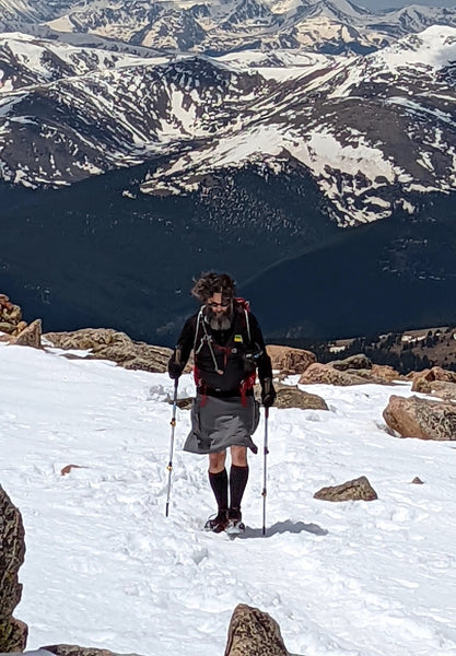 A bearded man hiking up a snowy mountain, with snowy mountains in the horizon. He has a greyish green skirt, hiking poles, long black socks, a black shirt, and a backpack.