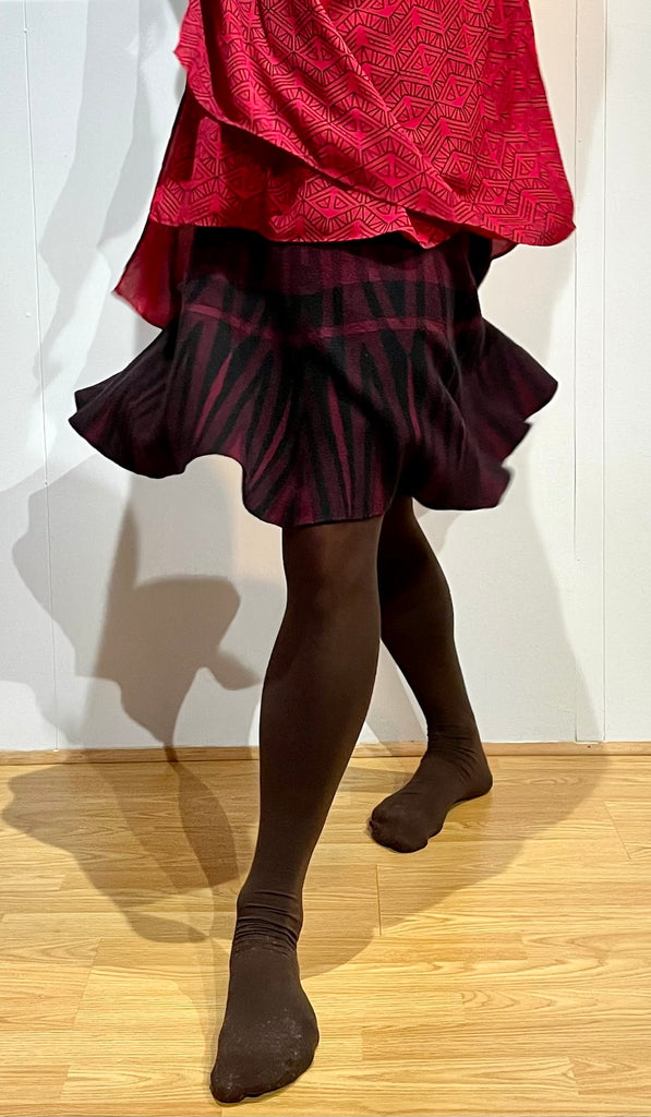 Male bodied person wearing a flowy, multi-layer red top with a black pattern, that extends below the waist, over a red and black thick wool skirt, with brown tights. Post is in a slight spin, with fabric dancing outward.