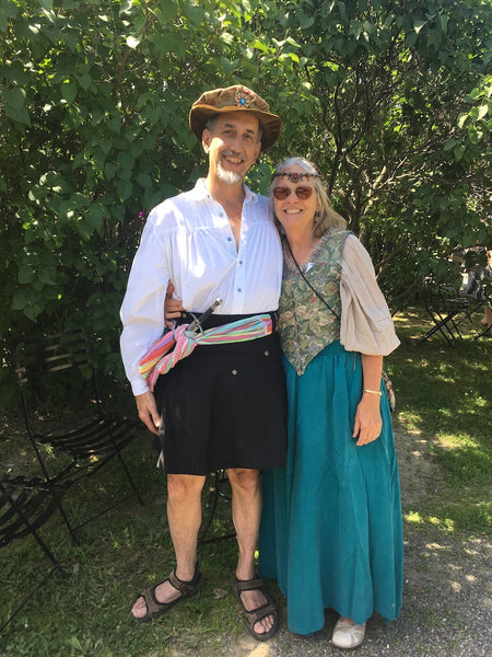 A couple posing together at Renn Faire, the man on the left in a black skirt, hat, and sword in a colourful cloth belt. A woman on the right, in a blue-green dress, flower pattern vest, and headband.