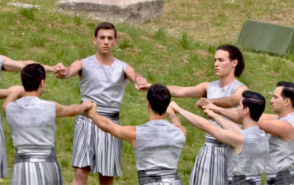 Male performers wear white and grey knee length skirts, on a grassy hill, holding hands in a circle.